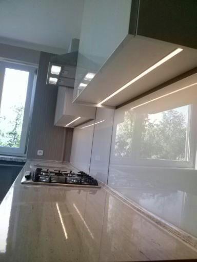 Jasna kuchnia z błyszczącymi frontami, oświetleniem LED podszafkowym i beżowym blatem z kuchenką gazową wbudowaną przy oknie.