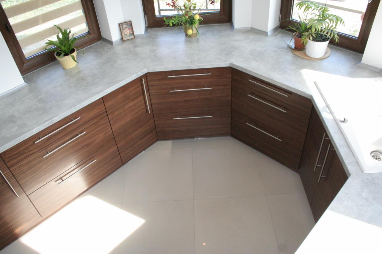 Widok z góry na narożną zabudowę kuchenną z ciemnego drewna z szarym blatem, z roślinami doniczkowymi przy oknach i jasną podłogą z płytek.