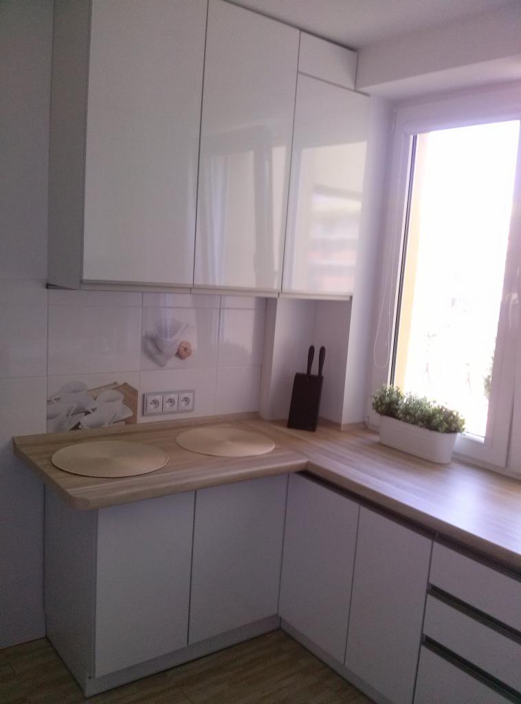 Jasna kuchnia z białymi szafkami, drewnianym blatem, okrągłymi podkładkami, kwiatami w doniczce na parapecie i dekoracyjnymi płytkami z motywem kawy.