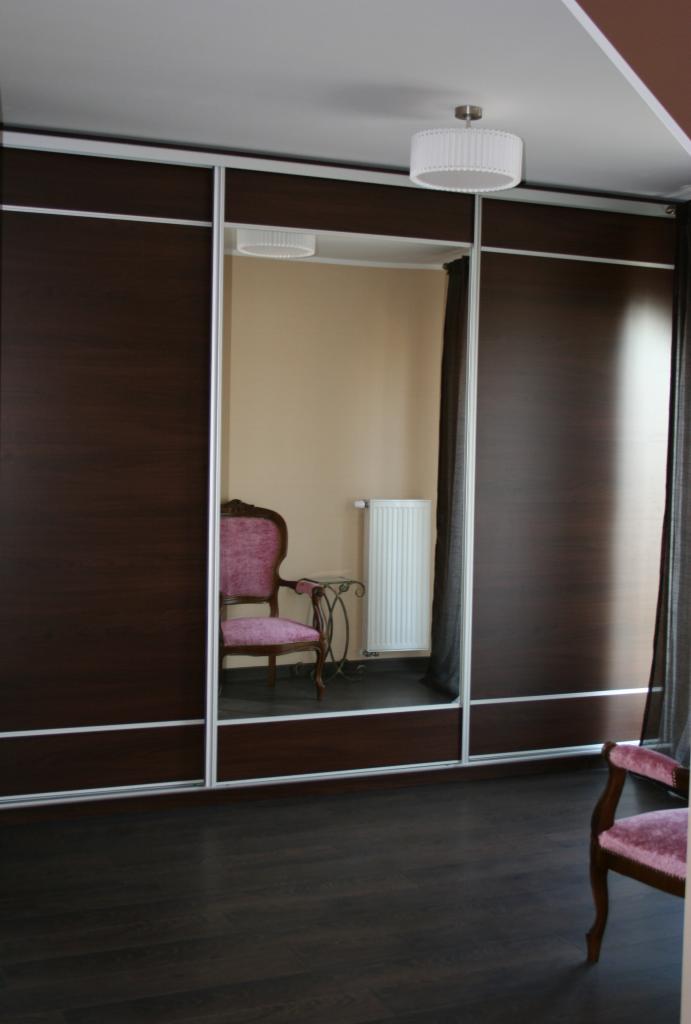 Ciemnobrązowa szafa przesuwna z białymi obramowaniami, odbicie w lustrze ukazuje krzesło w stylu vintage z fioletowym obiciem i grzejnik.