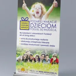 Roll-up reklamowy z wizerunkiem uśmiechniętej dziewczynki i grupy dzieci leżących na trawie, promujący akcję 'Podaruj Wakacje Dzieciom' Fundacji Art of Living, z widocznym adresem strony internetowej.
