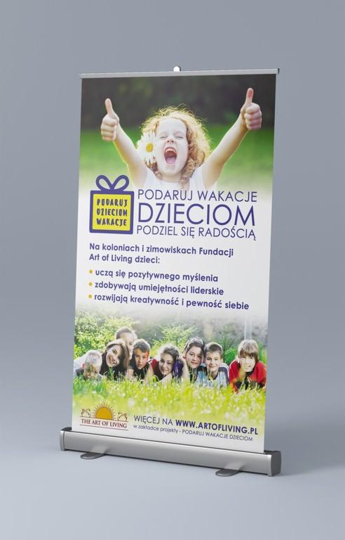 Roll-up reklamowy z wizerunkiem uśmiechniętej dziewczynki i grupy dzieci leżących na trawie, promujący akcję 'Podaruj Wakacje Dzieciom' Fundacji Art of Living, z widocznym adresem strony internetowej.
