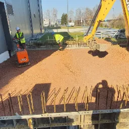 Przygotowanie podłoża pod budynek: ubijanie gruntu zagęszczarką wibrującą, zbrojenie wystające z szalunków, koparka JCB nasypująca ziemię, pracownicy w odblaskowych kamizelkach.