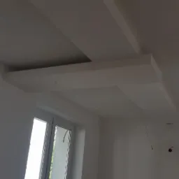 Biały sufit z geometrycznymi elementami i wnęką, widoczne wystające przewody elektryczne i okno