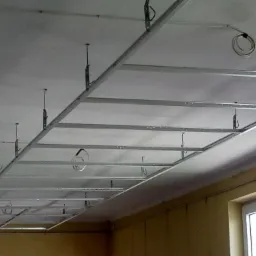 Metalowa konstrukcja sufitu podwieszanego z płyt kartonowo-gipsowych, widoczne przewody elektryczne, częściowo zamontowane płyty.