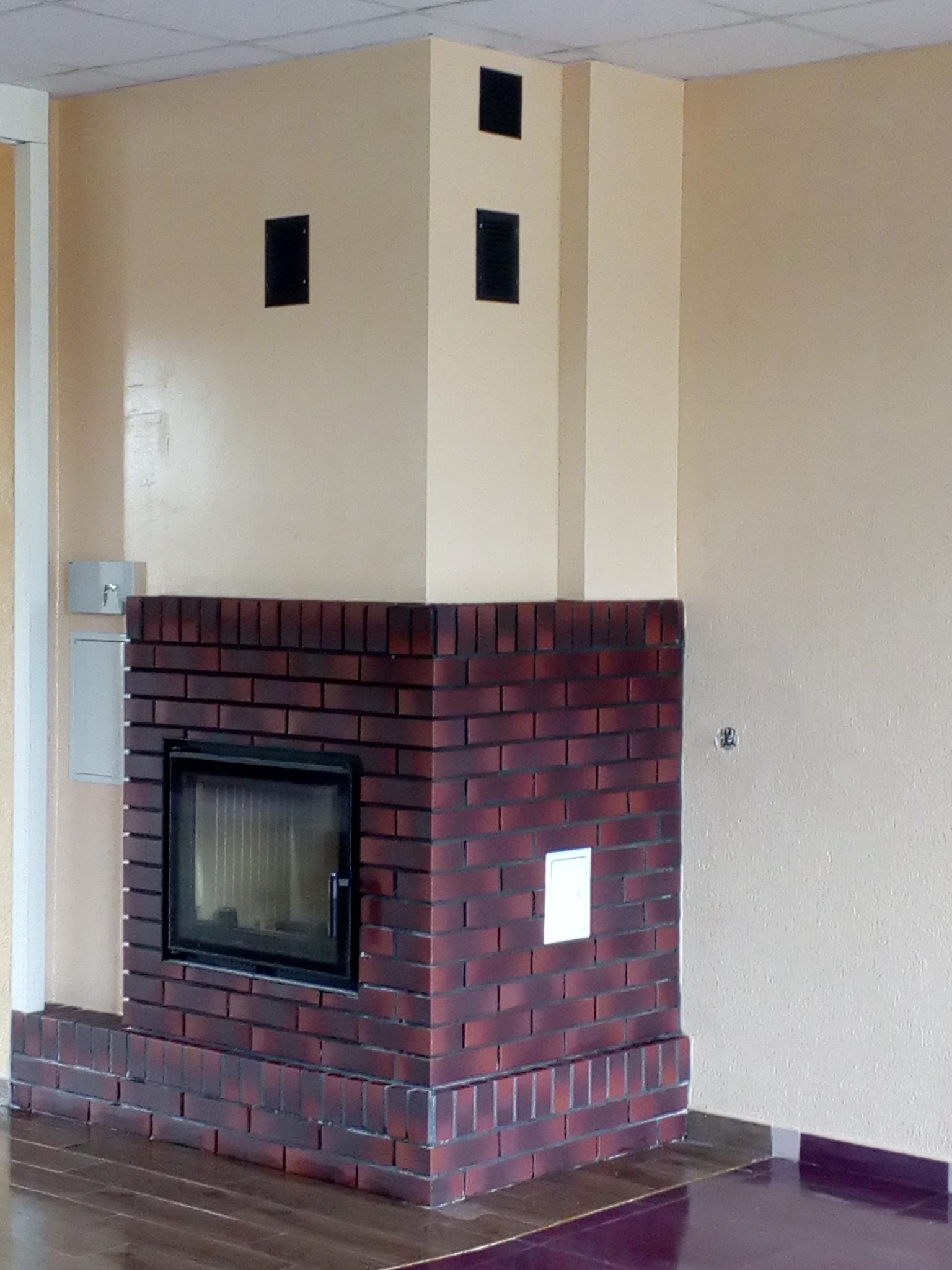 Murowany kominek z wkładem kominkowym, wykończony czerwoną cegłą, na tle beżowej ściany z trzema czarnymi otworami wentylacyjnymi, widoczna podłoga z ciemnego drewna.