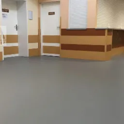 Szara, gładka podłoga w pomieszczeniu z beżowymi ścianami i brązowymi pasami ochronnymi, widoczne drzwi i okienko obsługi z roletą.