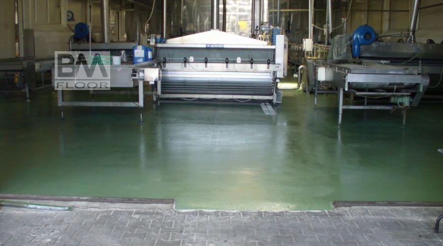 Zielona, błyszcząca podłoga w hali przemysłowej z widoczną linią produkcyjną i logo firmy BAA FLOOR w lewym górnym rogu.