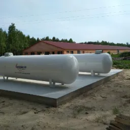 Dwa białe, poziome zbiorniki na gaz opałowy marki Gazgrod, umieszczone na betonowej platformie, z budynkiem z czerwonym dachem i lasem w tle.