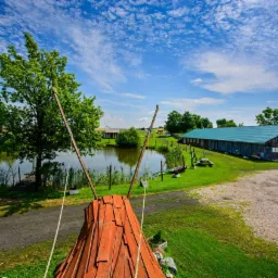 Widok na ośrodek rekreacyjny z drewnianym tipi na pierwszym planie, staw, budynek z zielonym dachem i błękitne niebo z chmurami w tle.