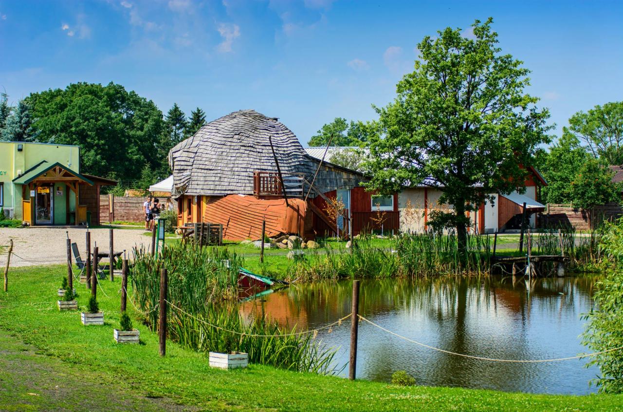 Kompleks budynków z nietypowym okrągłym budynkiem z drewnianym dachem, stawem i zielenią w słoneczny dzień.