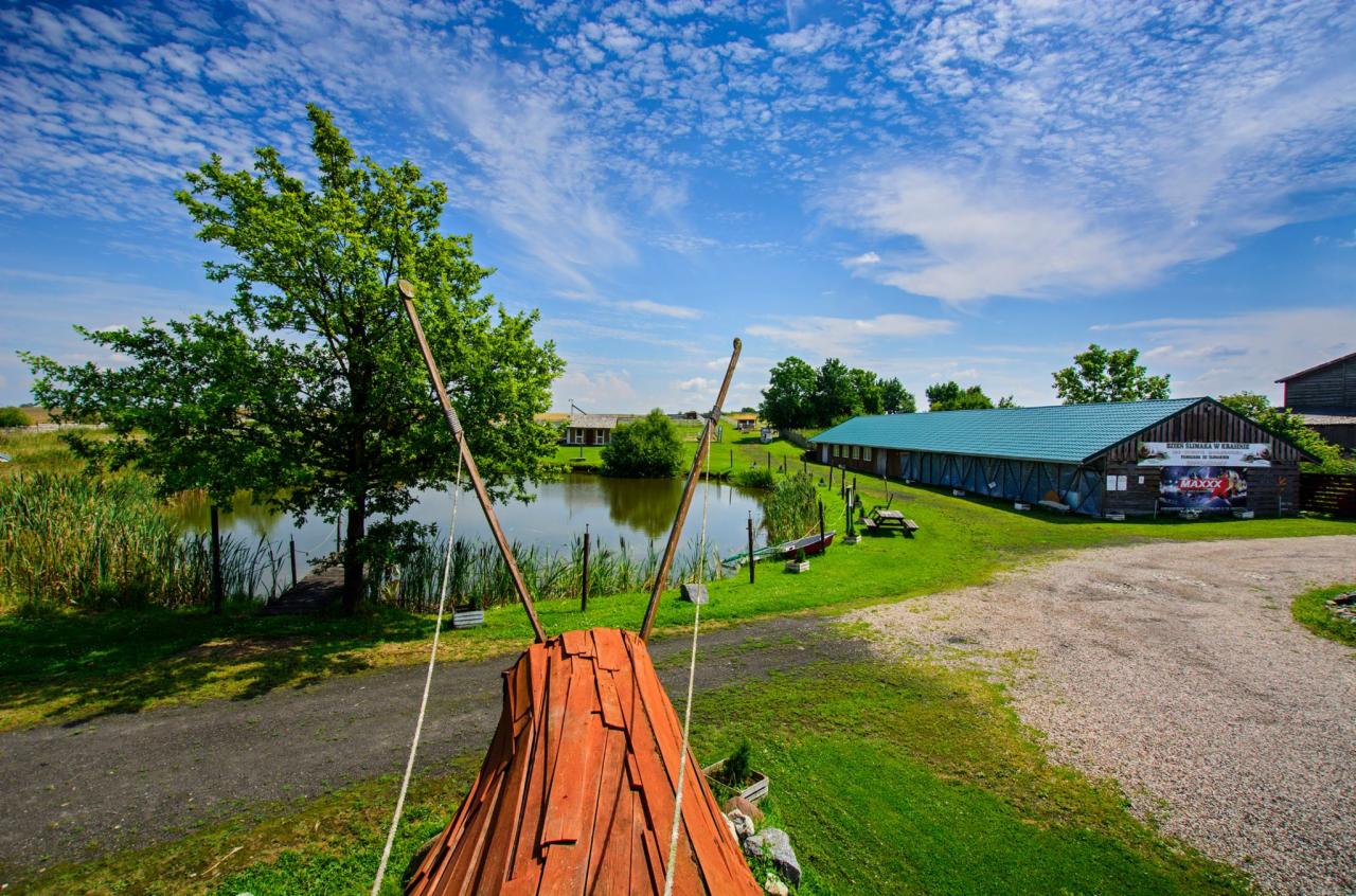 Widok na ośrodek rekreacyjny z drewnianym tipi na pierwszym planie, staw, budynek z zielonym dachem i błękitne niebo z chmurami w tle.
