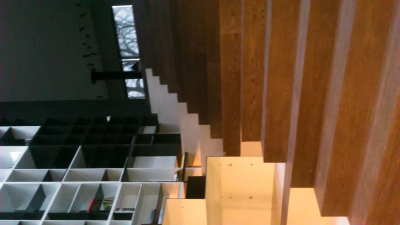 Drewniane schody z ażurową balustradą, widok z dołu na tle regału z półkami i okna.