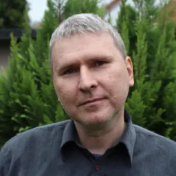 Portret mężczyzny z siwiejącymi włosami w ciemnej koszuli na tle zielonych krzew&oacute;w.