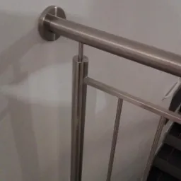 Fragment balustrady ze stali nierdzewnej, mocowanej do ściany, widok z góry na schody wyłożone ciemnymi płytkami.