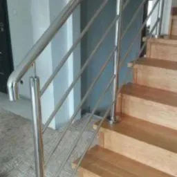 Nowoczesne schody drewniane z balustradą ze stali nierdzewnej, widok z dołu ukazujący drewniane stopnie i metalowe poręcze z wypełnieniem z cienkich prętów.