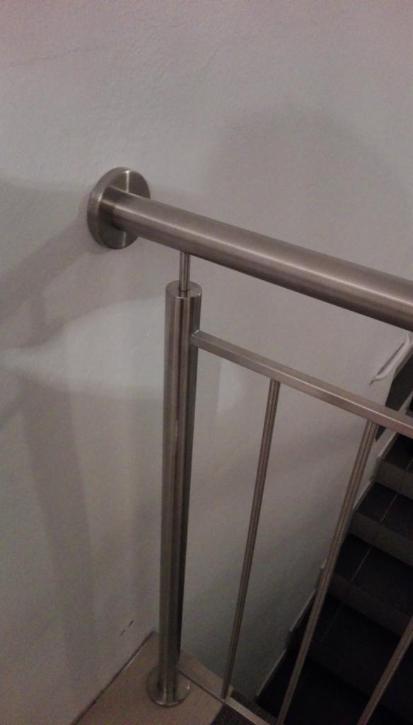 Fragment balustrady ze stali nierdzewnej, mocowanej do ściany, widok z góry na schody wyłożone ciemnymi płytkami.