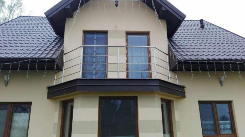 Dwukondygnacyjny dom z beżową elewacją, brązowym dachem i balkonem z prostą, metalową barierką, ozdobiony świątecznymi lampkami.