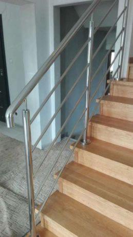 Nowoczesne schody drewniane z balustradą ze stali nierdzewnej, widok z dołu ukazujący drewniane stopnie i metalowe poręcze z wypełnieniem z cienkich prętów.