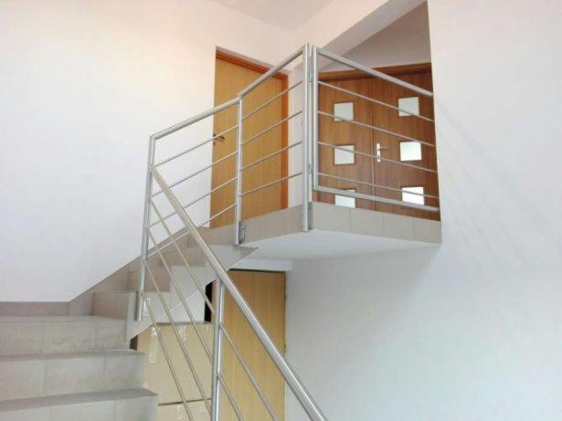 Schody z jasnymi stopniami i nowoczesną balustradą ze stali nierdzewnej, widok z dołu, minimalistyczne wnętrze z białymi ścianami i drewnianymi drzwiami w tle.