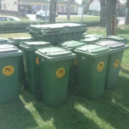 Stos zielonych kontenerów na kółkach z logo firmy LAD na trawie, jeden kontener umieszczony na górze pozostałych, w tle budynek i ogrodzenie.