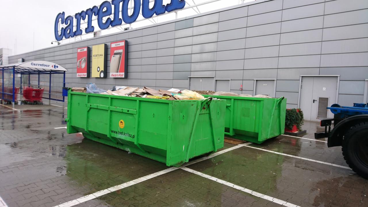 Dwa zielone, częściowo wypełnione kontenery na śmieci ustawione na parkingu przed sklepem wielkopowierzchniowym Carrefour w pochmurny dzień. Na kontenerze widoczna naklejka z adresem strony...