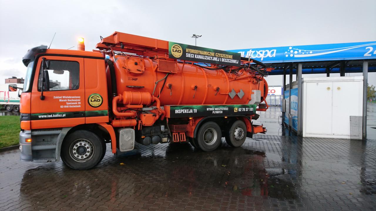 Pomarańczowa ciężarówka asenizacyjna firmy LAD z Kalisza, stojąca na mokrym placu przy stacji benzynowej Autospa, z widocznym napisem 'Hydrodynamiczne czyszczenie sieci kanalizacyjnej i deszczowej'...