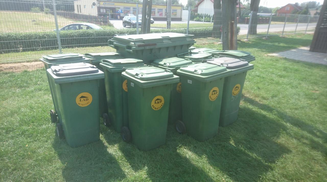 Stos zielonych kontenerów na kółkach z logo firmy LAD na trawie, jeden kontener umieszczony na górze pozostałych, w tle budynek i ogrodzenie.