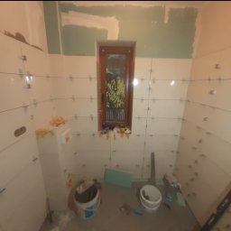 Wega Budownictwo - Łazienka w trakcie kładzenia białych płytek, widoczne krzyżyki dystansowe i poziomica. Okno z widokiem na zieleń. Częściowo położone płytki, etap prac wykończeniowych.