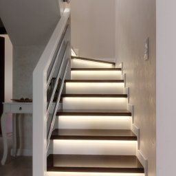 Wega Budownictwo - Nowoczesne schody z podświetleniem LED na stopniach, białe balustrady z metalowymi elementami. Minimalistyczny design, jasne ściany z delikatną fakturą.