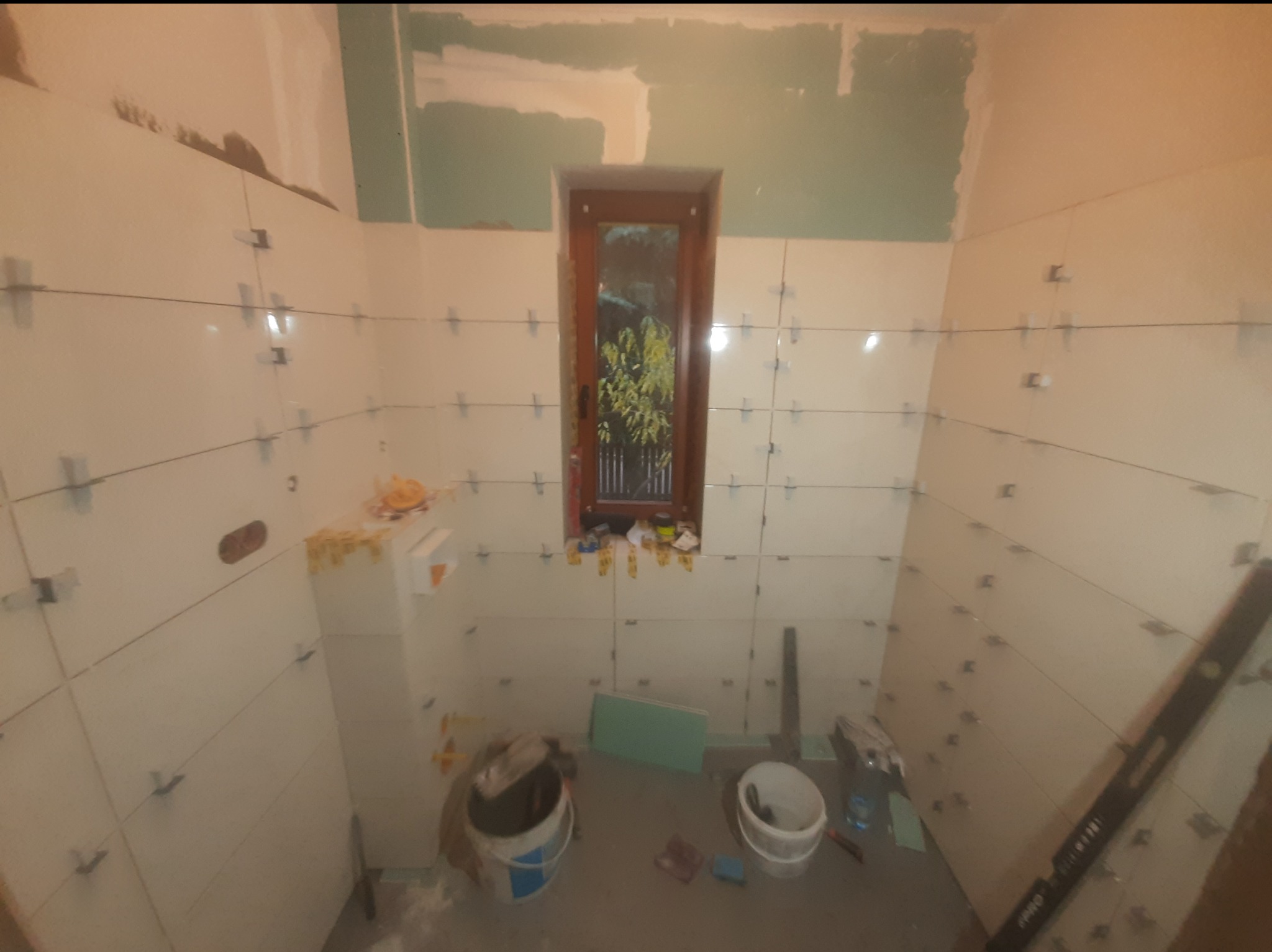 Łazienka w trakcie kładzenia białych płytek, widoczne krzyżyki dystansowe i poziomica. Okno z widokiem na zieleń. Częściowo położone płytki, etap prac wykończeniowych.