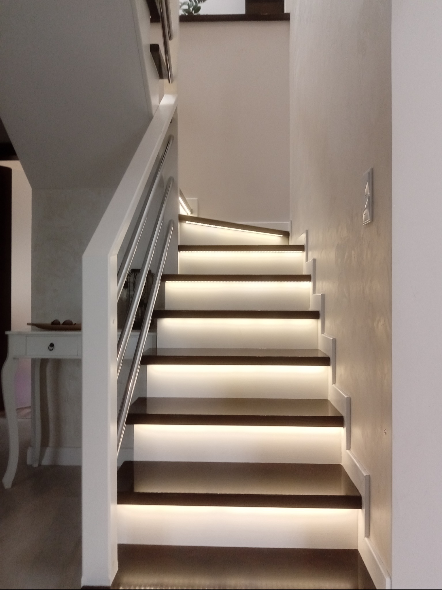 Nowoczesne schody z podświetleniem LED na stopniach, białe balustrady z metalowymi elementami. Minimalistyczny design, jasne ściany z delikatną fakturą.