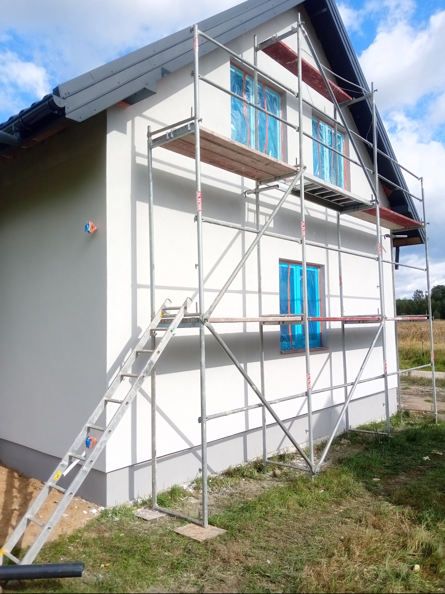 Biały dom jednorodzinny z szarym dachem, otoczony rusztowaniem. Okna zabezpieczone folią. Świeżo otynkowana elewacja w słoneczny dzień.