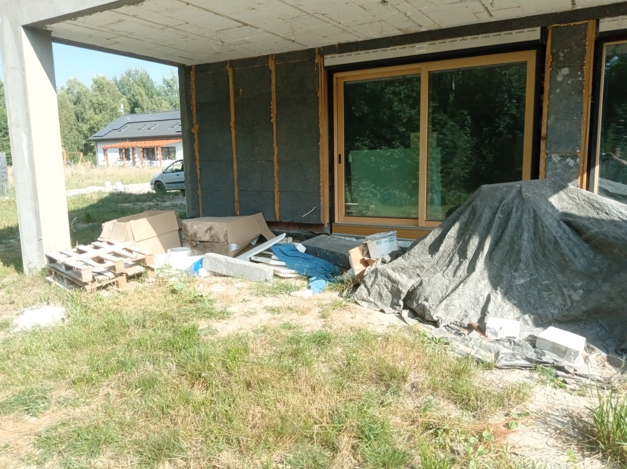 Budowa domu w trakcie ocieplania: widoczne płyty izolacyjne na ścianach, okna z drewnianą ramą, palety i materiały budowlane na trawie. Dom w tle.