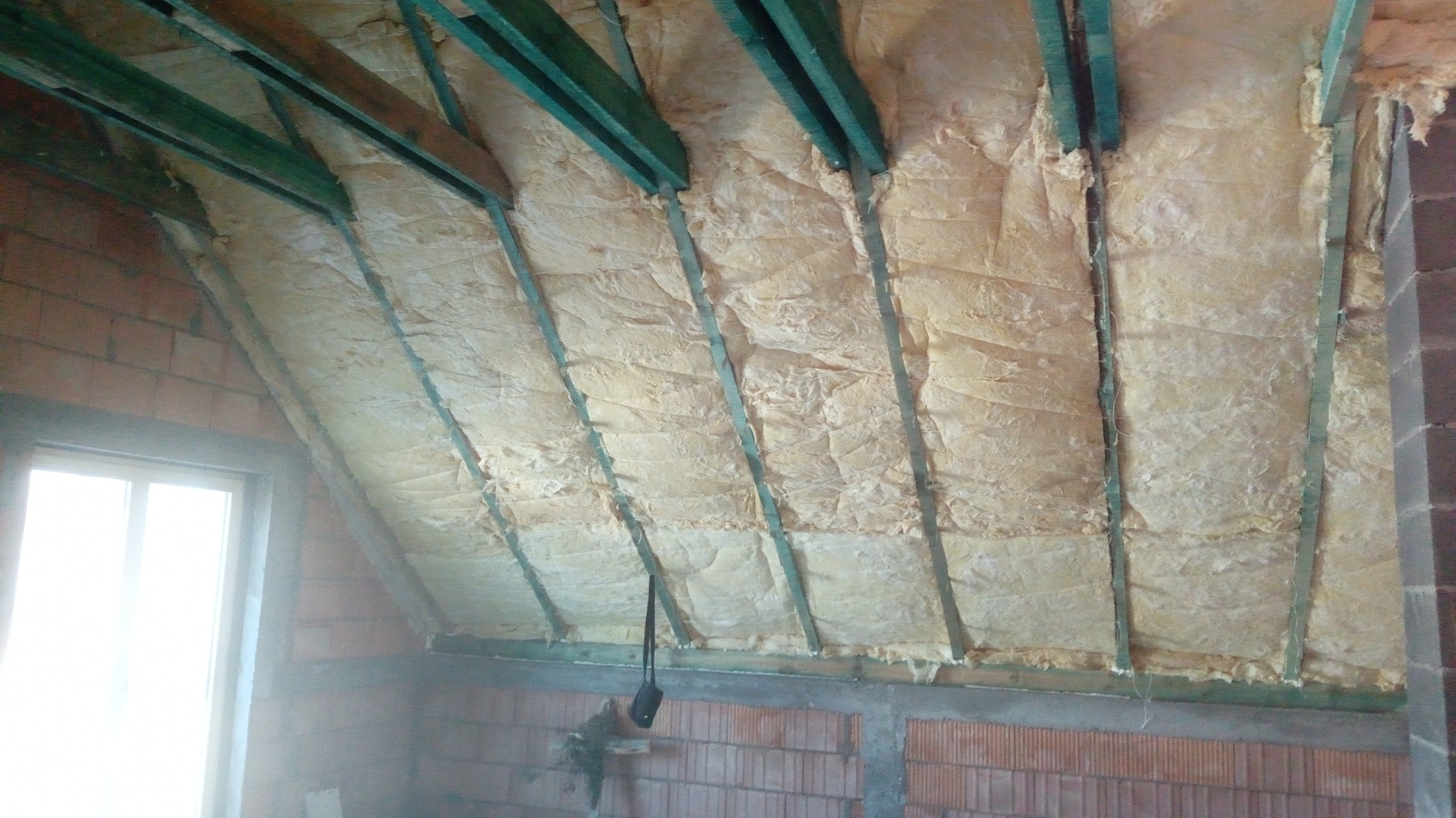 Izolacja poddasza wełną mineralną między zielonymi krokwiami, widoczne ceglane ściany i okno. Etap prac wykończeniowych w budynku mieszkalnym.