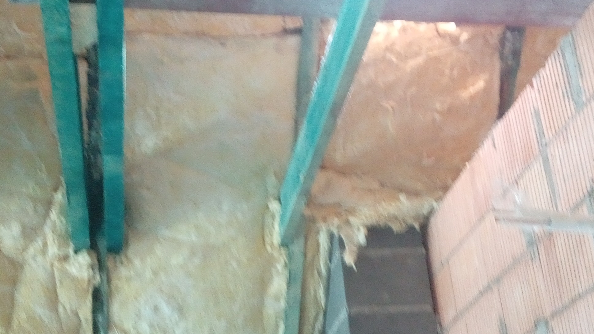 Izolacja poddasza wełną mineralną, widoczne drewniane elementy konstrukcyjne i fragment ściany z cegły. Ujęcie z dołu, podkreślające detale wykonania.