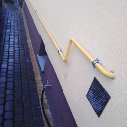 Hydro-Invest Łukasz Nowak - Żółta rura gazowa przymocowana do elewacji budynku z widocznym zaworem gazowym i fragmentem chodnika z kostki brukowej