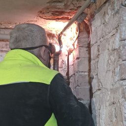 Hydro-Invest Łukasz Nowak - Spawanie rur w trudnych warunkach: hydraulik w okularach spawalniczych łączy rury w wąskiej przestrzeni, widoczne iskry i surowa ceglana ściana.