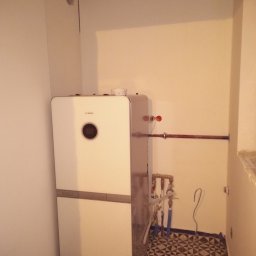 Hydro-Invest Łukasz Nowak - Nowoczesny, biały piec gazowy Bosch zamontowany w pomieszczeniu z wzorzystą, czarno-białą podłogą. Widoczne rury miedziane i przyłącza hydrauliczne. Na ścianie widoczne ślady po instalacji.
