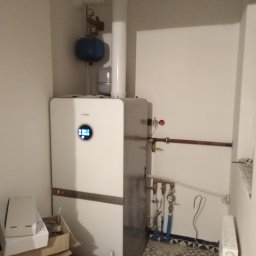 Hydro-Invest Łukasz Nowak - Biały, nowoczesny piec gazowy marki Bosch zamontowany w rogu pomieszczenia, z widocznymi rurami, manometrem i zbiornikiem wyrównawczym. Na podłodze wzorzysta mozaika. Obok kartony po akcesoriach.