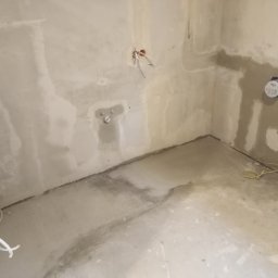 Hydro-Invest Łukasz Nowak - Surowe wnętrze łazienki w trakcie remontu, widoczne wystające kable, rury i niedokończone ściany z szarym tynkiem, przygotowane pod instalacje hydrauliczne.