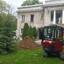 Czerwono-czarna mini koparka Yanmar na trawniku przed budynkiem z kolumnami i stertą ziemi, widoczny numer telefonu na kabinie.