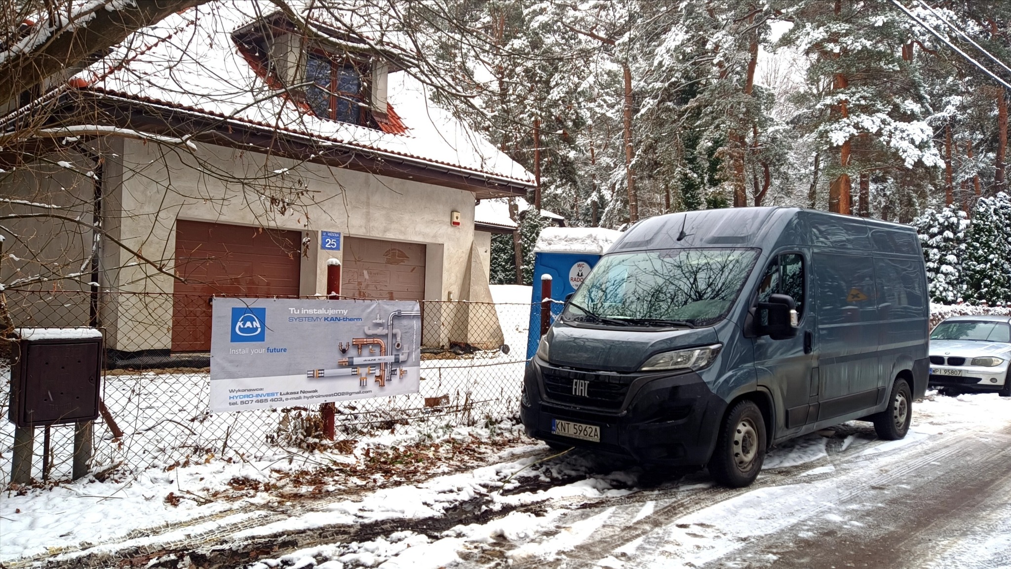 Zimowy krajobraz z busem firmy instalacyjnej na tle domu z banerem reklamowym systemów KAN-therm. Widoczny przenośny WC i zaparkowany samochód osobowy.