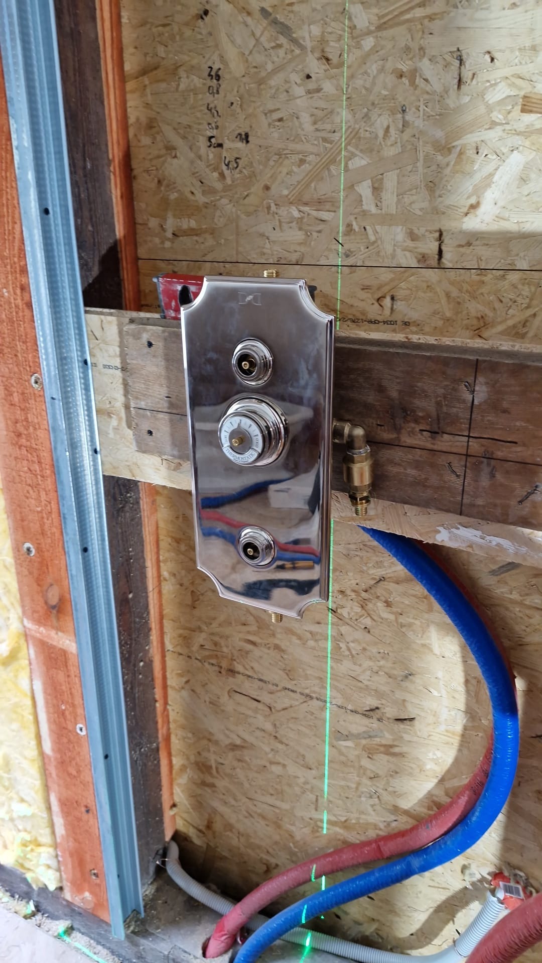 Błyszcząca, chromowana bateria termostatyczna zamontowana na drewnianej ścianie w trakcie instalacji, z widocznymi rurami PEX i poziomnicą laserową.