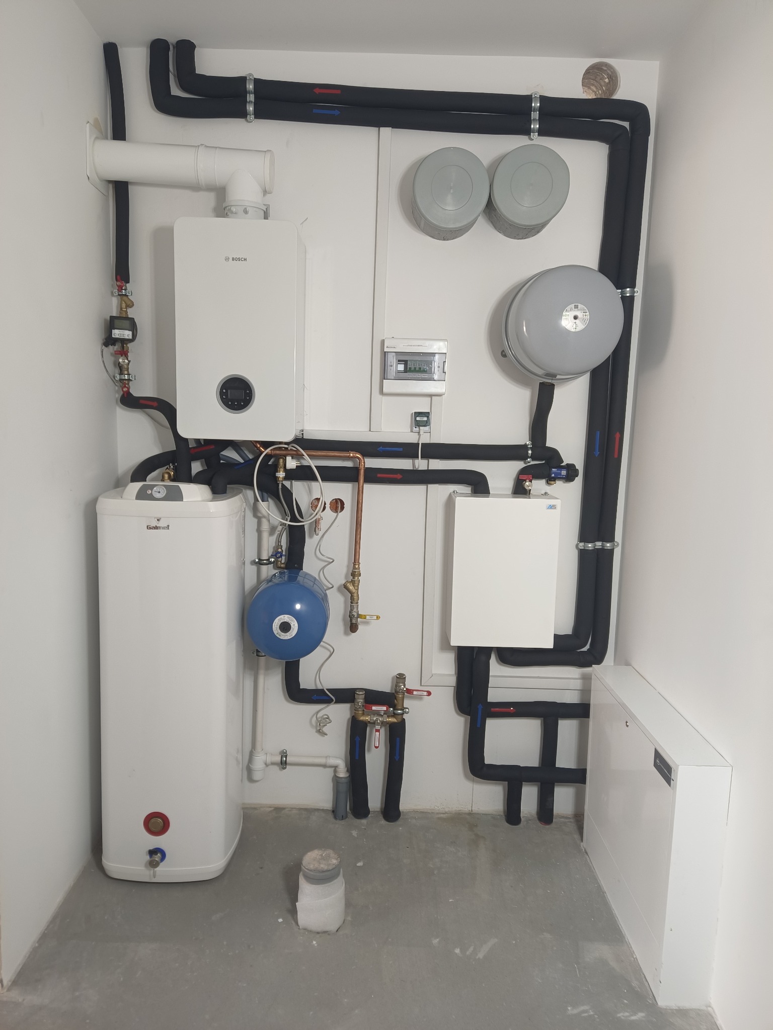 Nowoczesna kotłownia z białym piecem gazowym Bosch, zasobnikiem Galmet i czarnymi rurami w izolacji. Widoczne elementy instalacji CO i CWU, szara podłoga.
