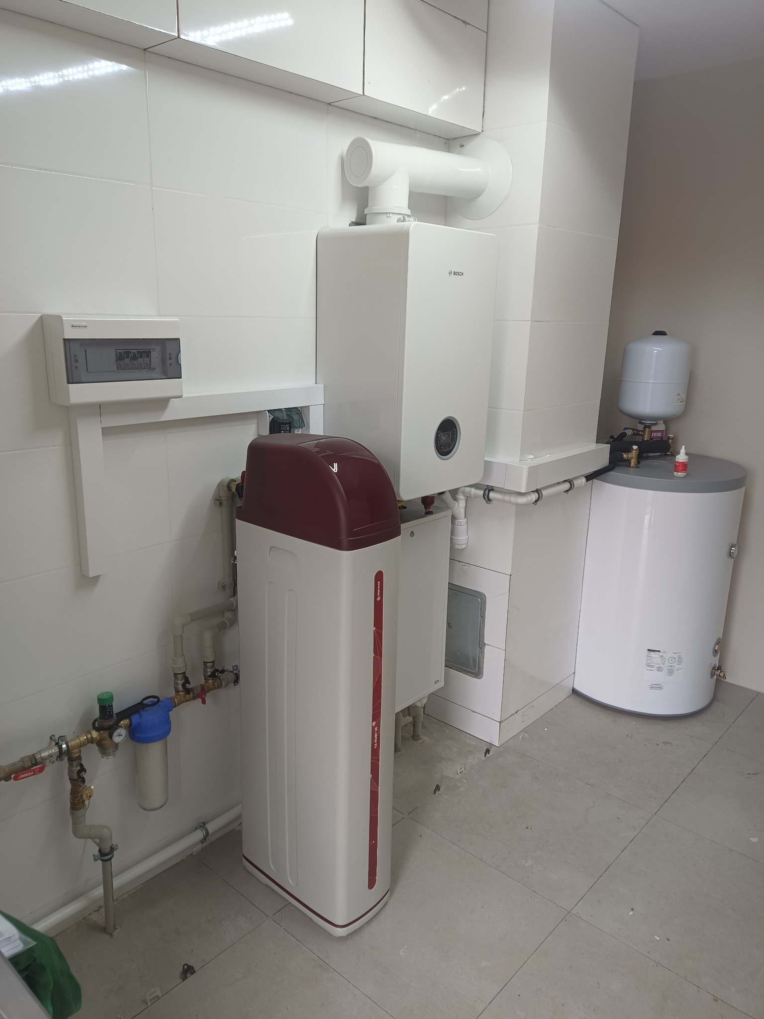 Biała kotłownia z piecem gazowym Bosch, zasobnikiem wody i zmiękczaczem wody z czerwoną głowicą. Widoczne rury, filtry i płytki ceramiczne na ścianach i podłodze.