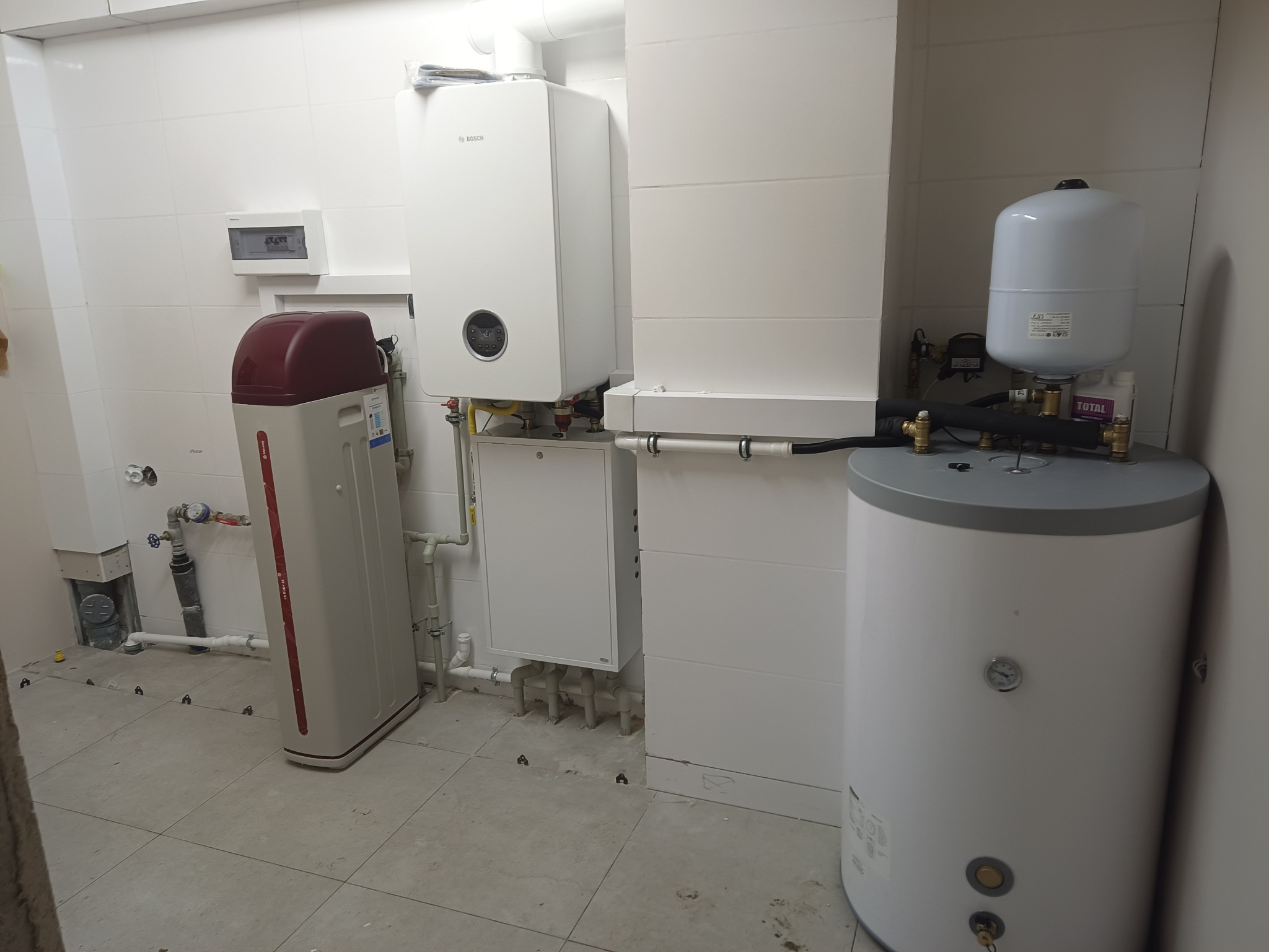 Białe pomieszczenie z zainstalowanym kotłem gazowym Bosch, zasobnikiem ciepłej wody i zmiękczaczem wody. Widoczne rury i elementy instalacji. Ułożone płytki na podłodze.