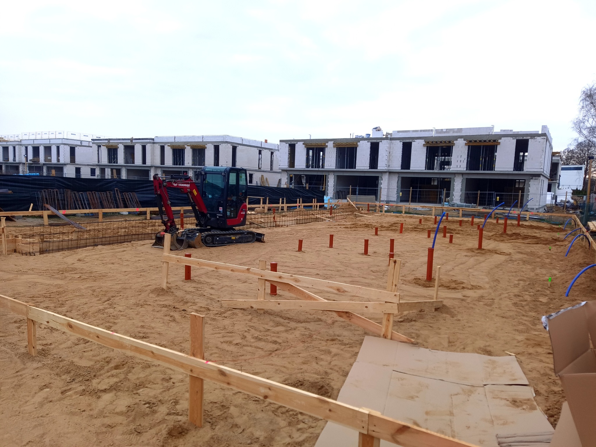 Fundamenty pod budowę szeregowców z widoczną instalacją hydrauliczną i mini koparką Yanmar na piaszczystym terenie. Ujęcie z poziomu gruntu, pochmurne niebo.