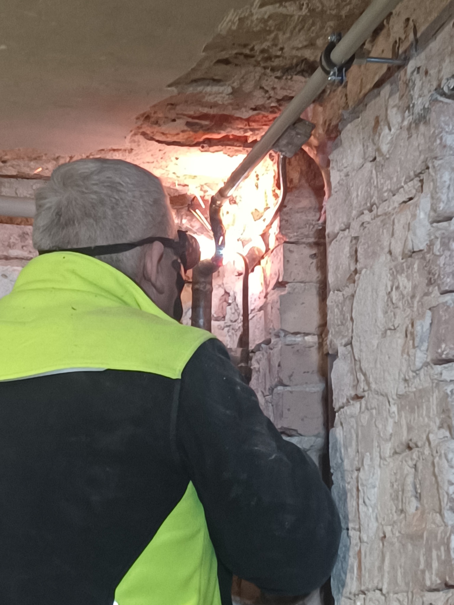 Spawanie rur w trudnych warunkach: hydraulik w okularach spawalniczych łączy rury w wąskiej przestrzeni, widoczne iskry i surowa ceglana ściana.