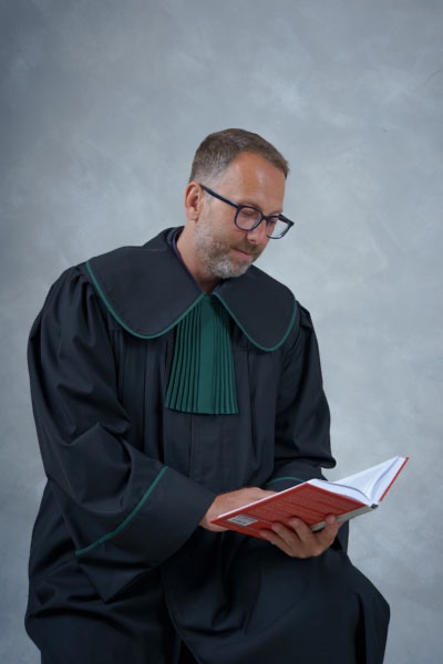 Adwokat w todze z zielonym żabotem, w okularach, czyta książkę z czerwoną okładką na rozmytym szarym tle. Ujęcie od pasa w górę, z delikatnym uśmiechem.